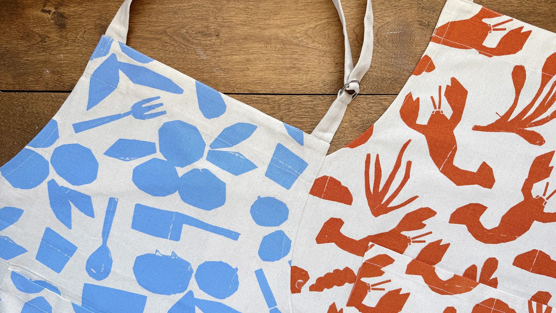 Two cotton aprons with bold, screen-printed designs laid flat on a wooden surface. The left apron features a blue pattern of kitchen utensils and blueberries, while the right apron showcases an orange lobster and seaweed motif. Both have adjustable neck straps and a natural off-white fabric base.