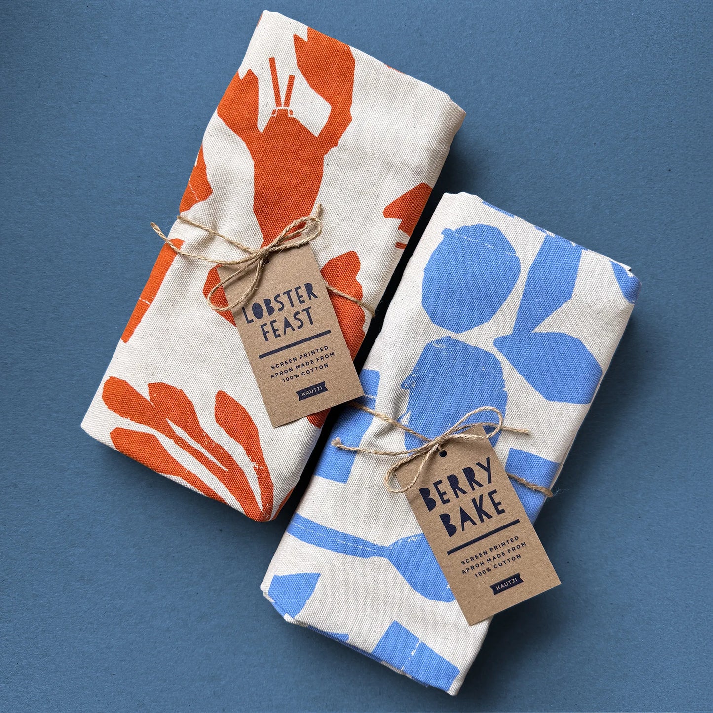 The Lobster Feast and Berry Bake aprons side by side, both folded and tied with twine, showing their distinct red and blue patterns.