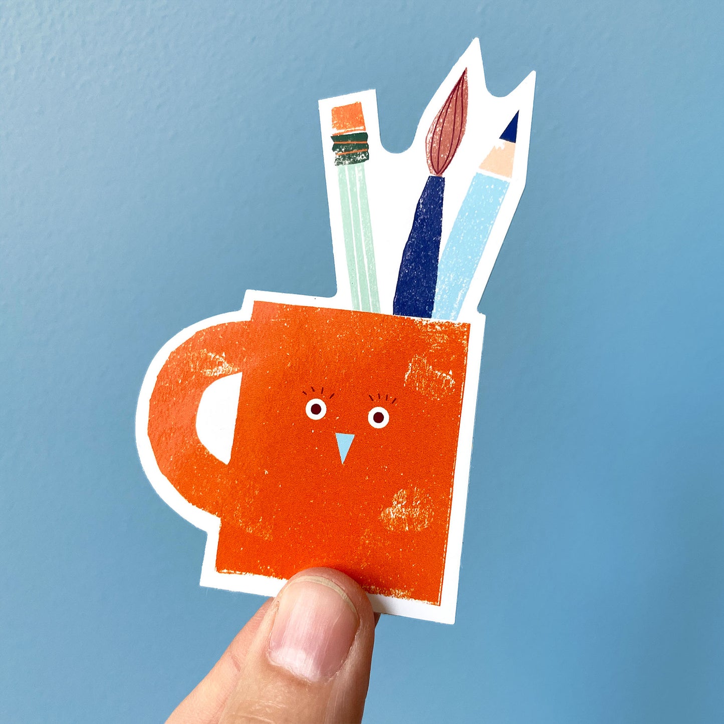Cute orange pencil mug sticker with art supplies, paintbrush and pencils sticking out of a playful bird-shaped holder