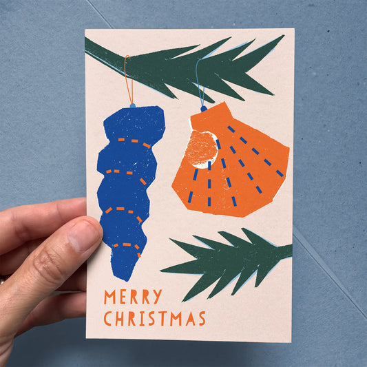 Hand holding a Christmas card with blue and orange ornaments and 'Merry Christmas' text on a beige background.