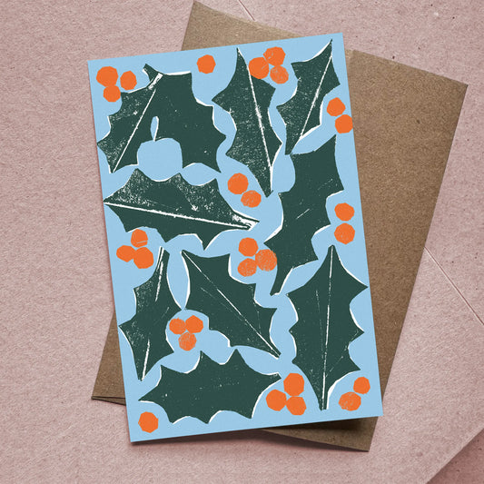 Flat Christmas card with holly leaves and berries illustration on blue background, paired with kraft envelope.