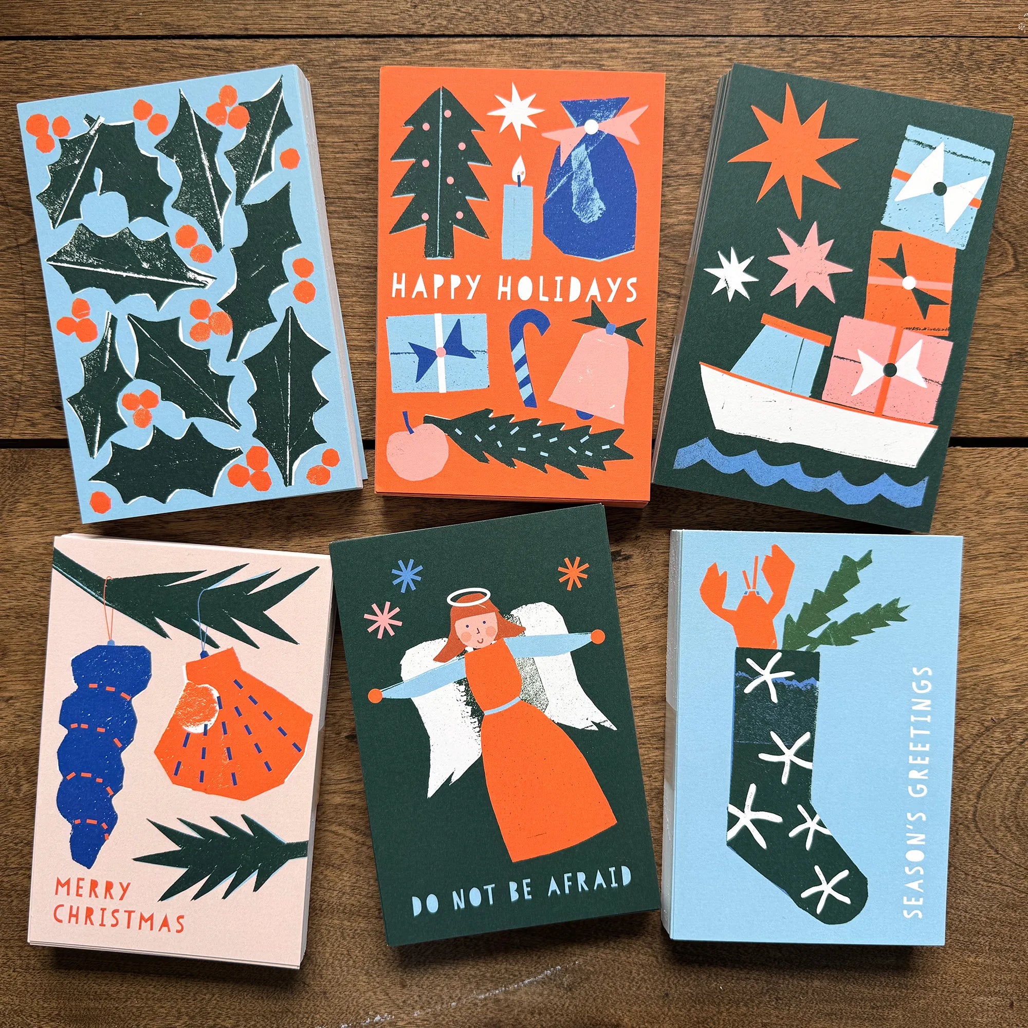 A set of six colourful flat Christmas cards by Kautzi Illustrations, designed in Cape Breton, Nova Scotia. The cards feature bold, paper-cut designs inspired by the Maritimes, including holly and berries, a lobster in a stocking, an angel, ornaments, a festive still life, and a boat with presents.