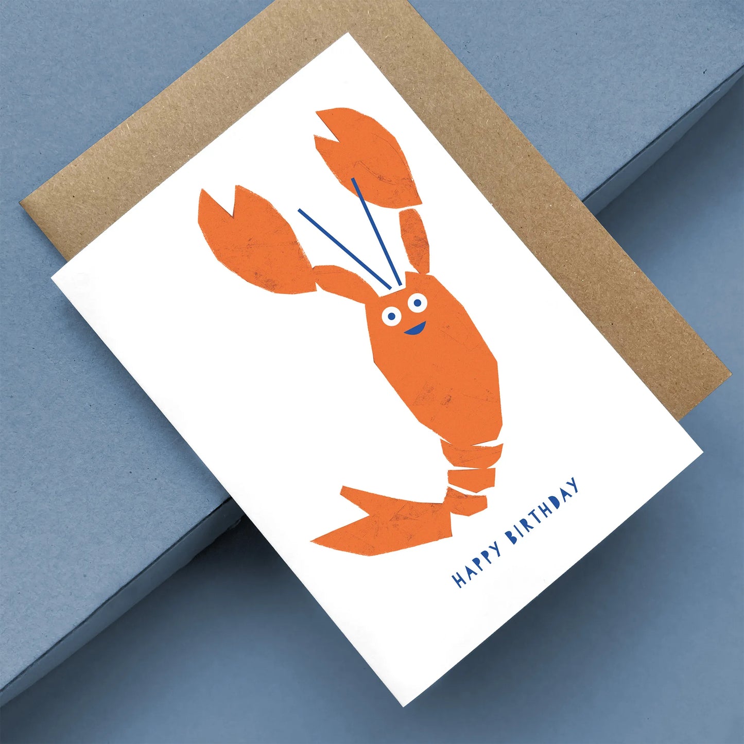 Kautzi greeting card featuring an orange lobster illustration with blue details and “Happy Birthday” text, shown at an angle on a blue background with a kraft envelope – designed in Cape Breton, Nova Scotia.