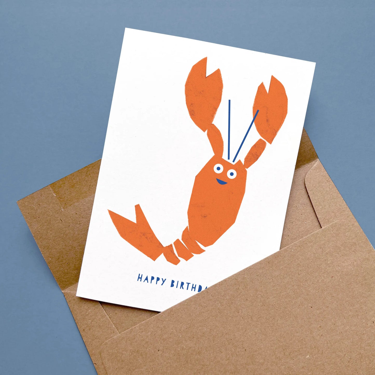 Kautzi lobster birthday card with orange lobster illustration and blue lettering, displayed flat on a blue background with a partially opened kraft envelope – designed in Cape Breton, Nova Scotia.