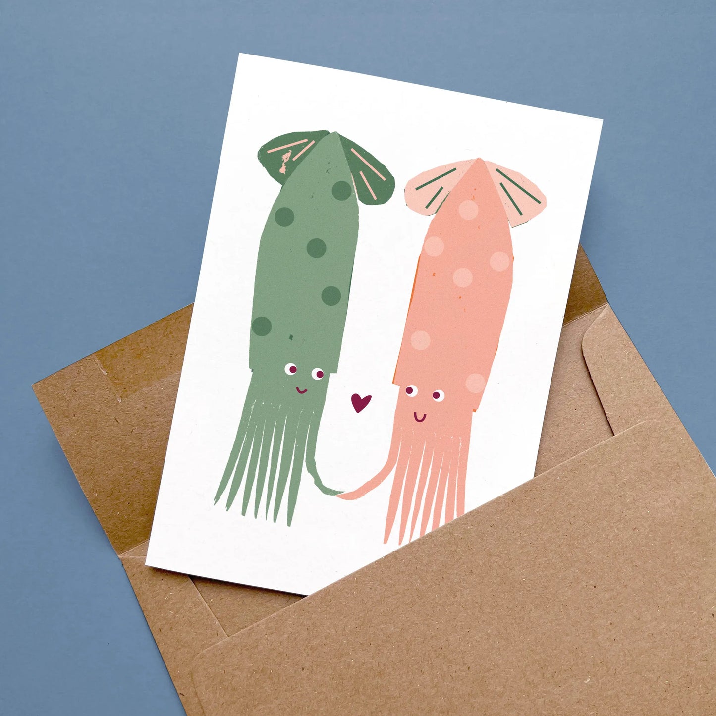 Kautzi squid greeting card with two playful squids in green and coral pink holding tentacles, displayed flat with a kraft envelope on a blue surface – designed in Cape Breton, Nova Scotia.