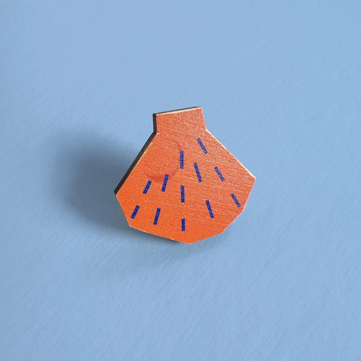 Close-up of Kautzi maple wood veneer shell pin badge in bright orange with blue lines, inspired by Nova Scotia shoreline finds