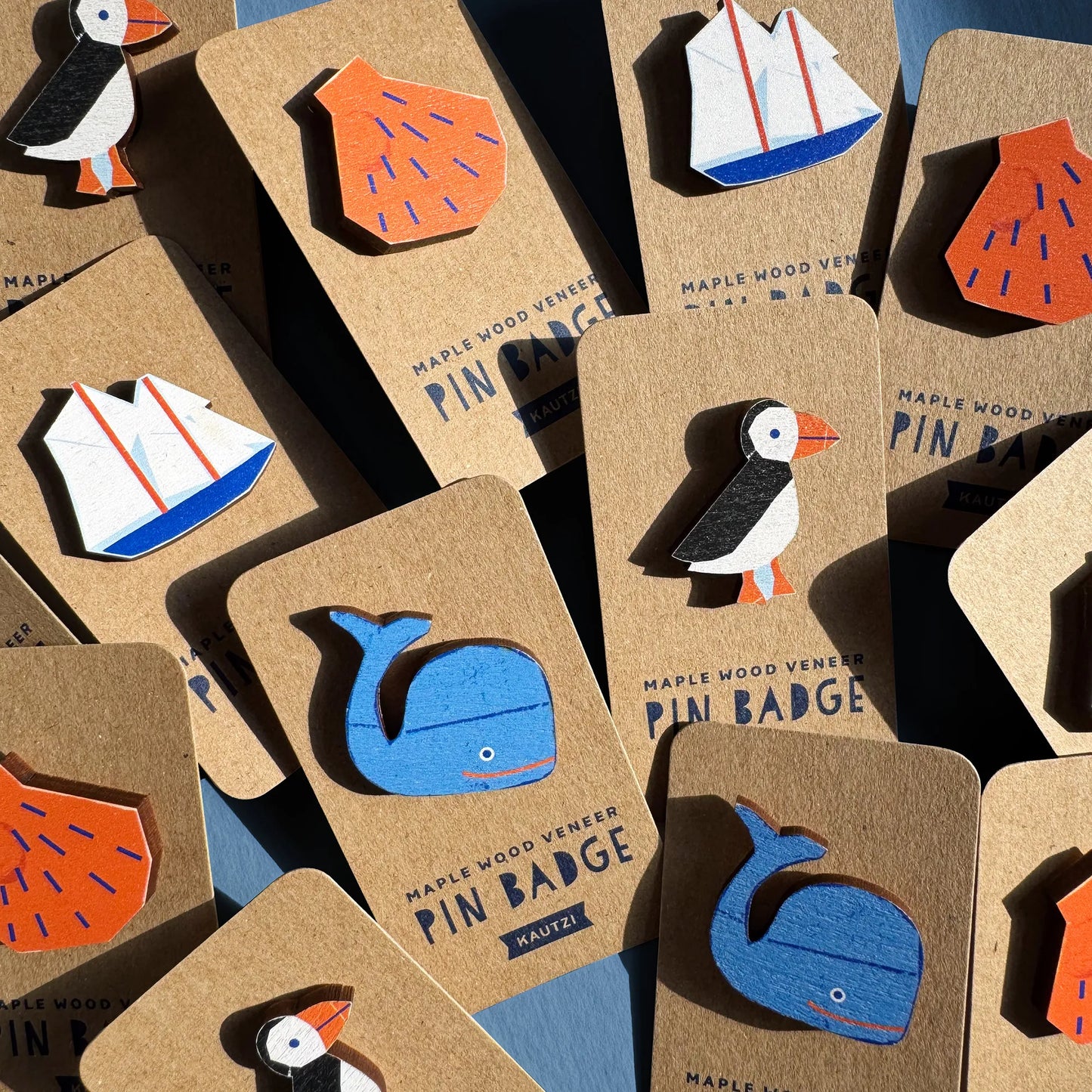 Assorted Kautzi maple wood veneer pin badges displayed on kraft backing cards, featuring puffin, whale, sailboat, and orange shell designs in bold, colourful prints.