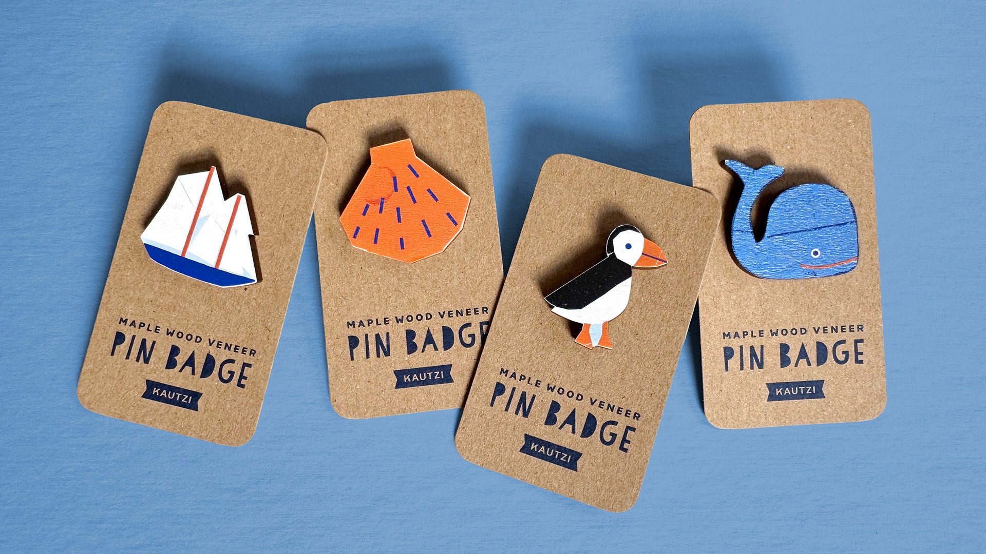 Four maple wood veneer pins designed by Kautzi displayed on kraft backing cards. The pins include a red and white sailboat, an orange shell with blue lines, a black and white puffin with an orange beak and feet, and a smiling blue whale. The cards are printed with "Maple Wood Veneer Pin Badge – Kautzi" in navy blue on a blue background.
