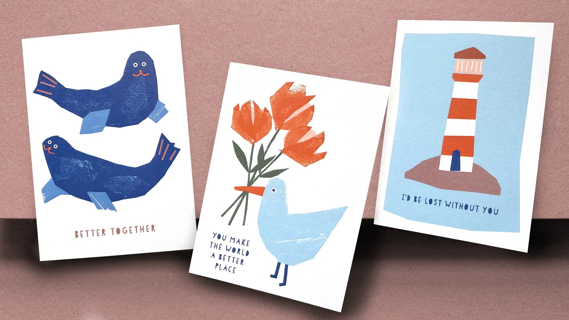 Three illustrated greeting cards laid out on a soft pinkish-brown background. The left card features two navy blue seals with small orange details, facing each other with the text "Better Together." The middle card shows a light blue seagull holding a bouquet of orange flowers with the text "You Make the World a Better Place." The right card depicts a red and white striped lighthouse on a small island, set against a blue sky, with the text "I'd Be Lost Without You."