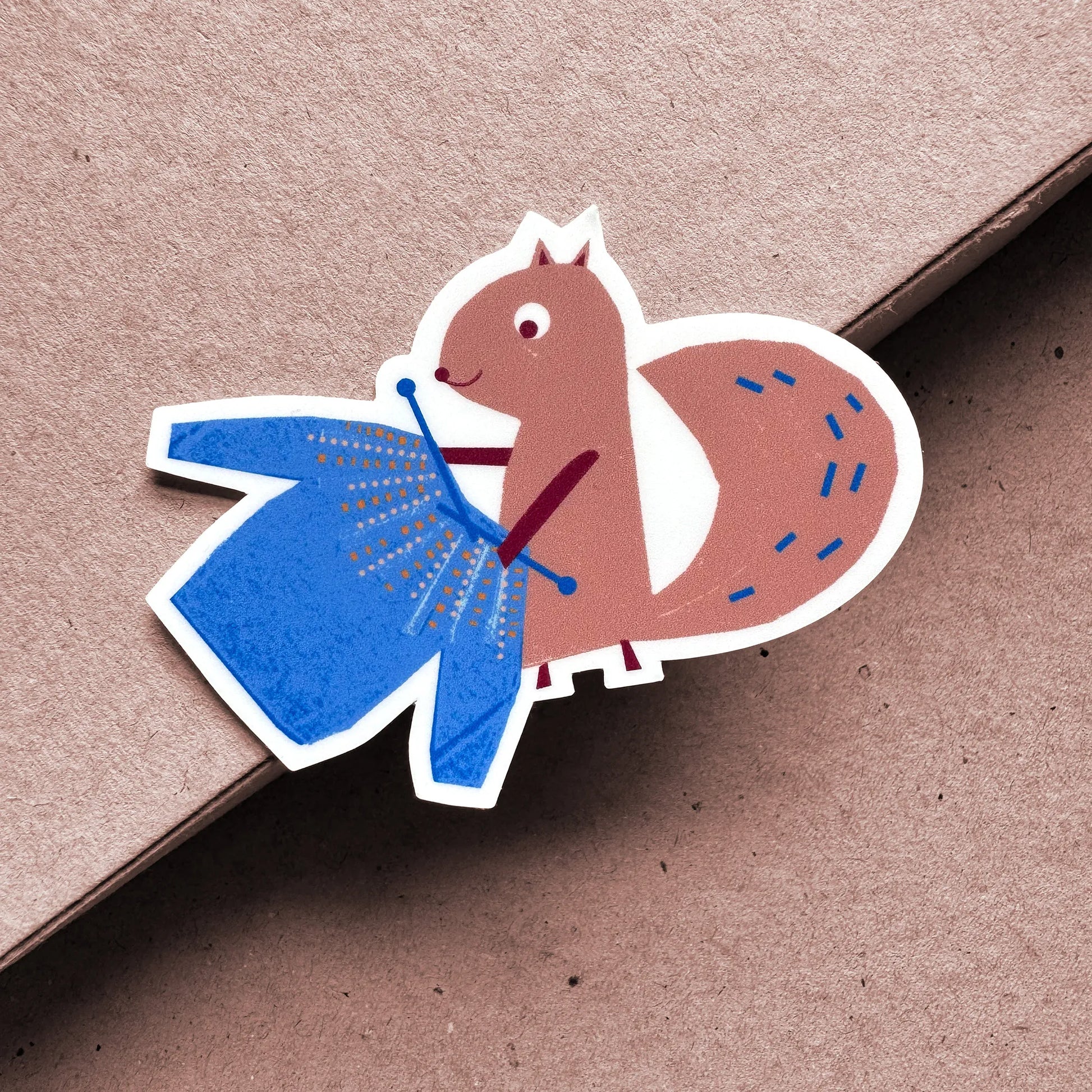 Full view of Kautzi Knitting Squirrel sticker on kraft background – illustrated squirrel knitting in blue, designed in Cape Breton, Nova Scotia.