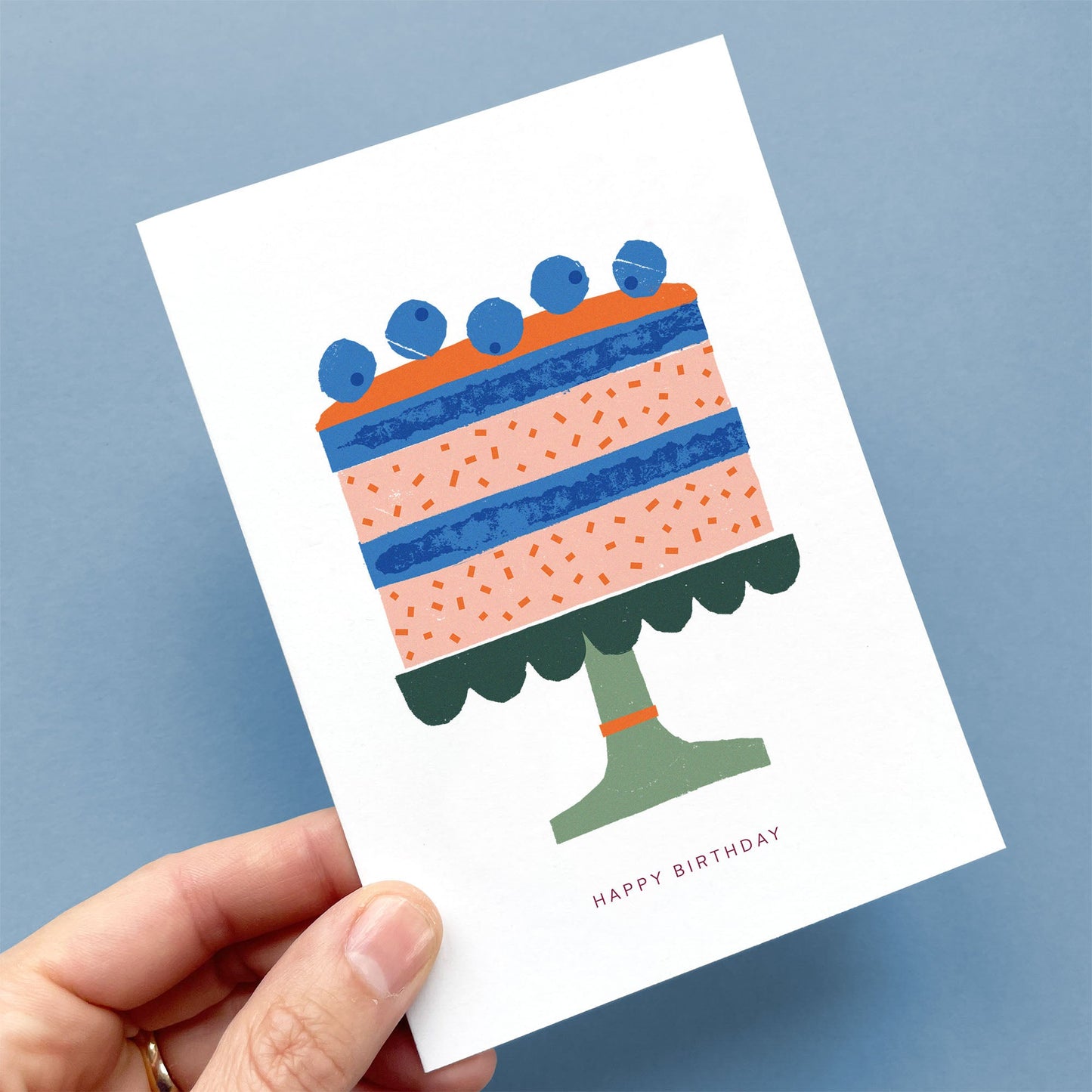 Greeting Card Birthday Cake featuring blueberry-topped layered cake in coral and blue on green cake stand, illustrated with cardboard