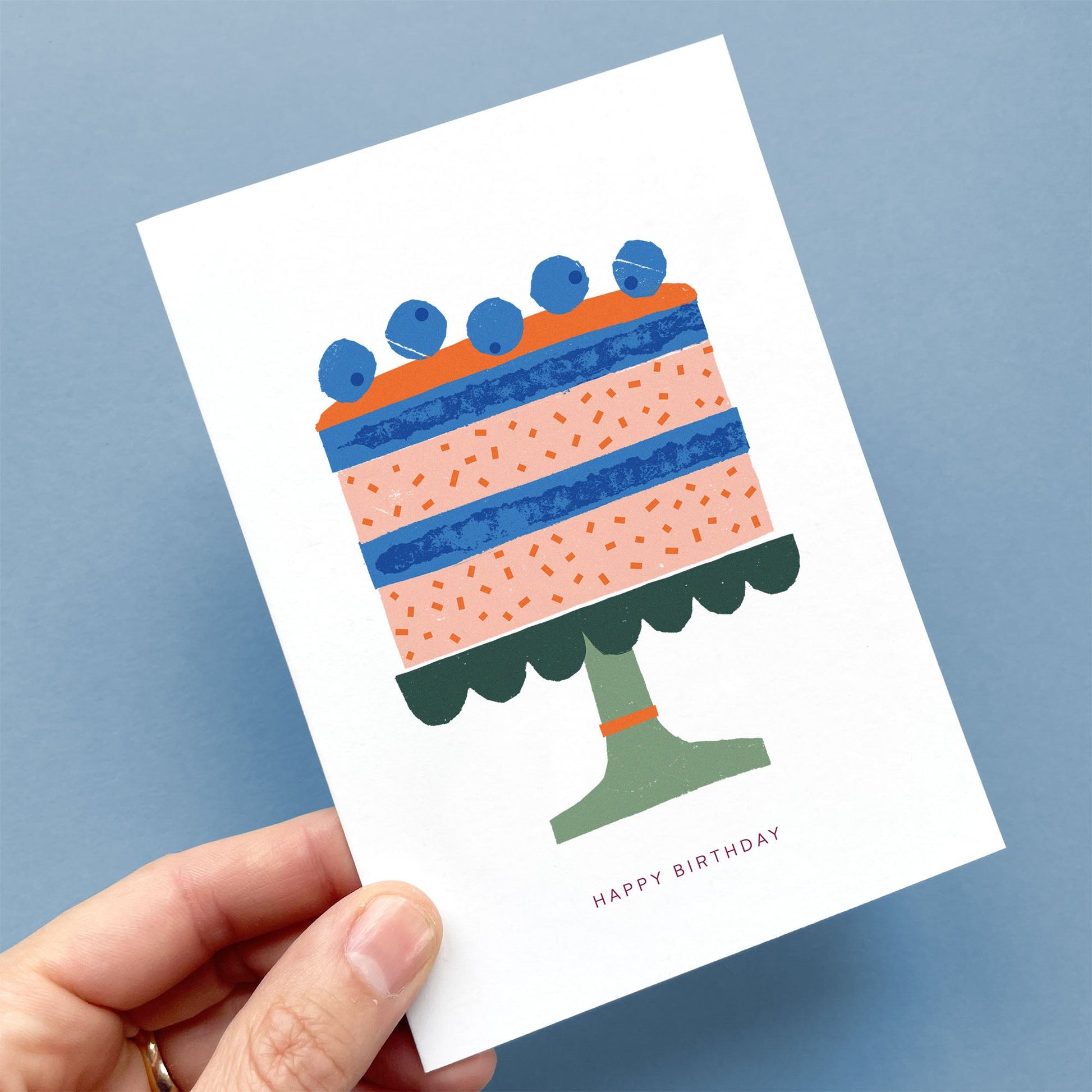 Greeting Card Birthday Cake featuring blueberry-topped layered cake in coral and blue on green cake stand, illustrated with cardboard
