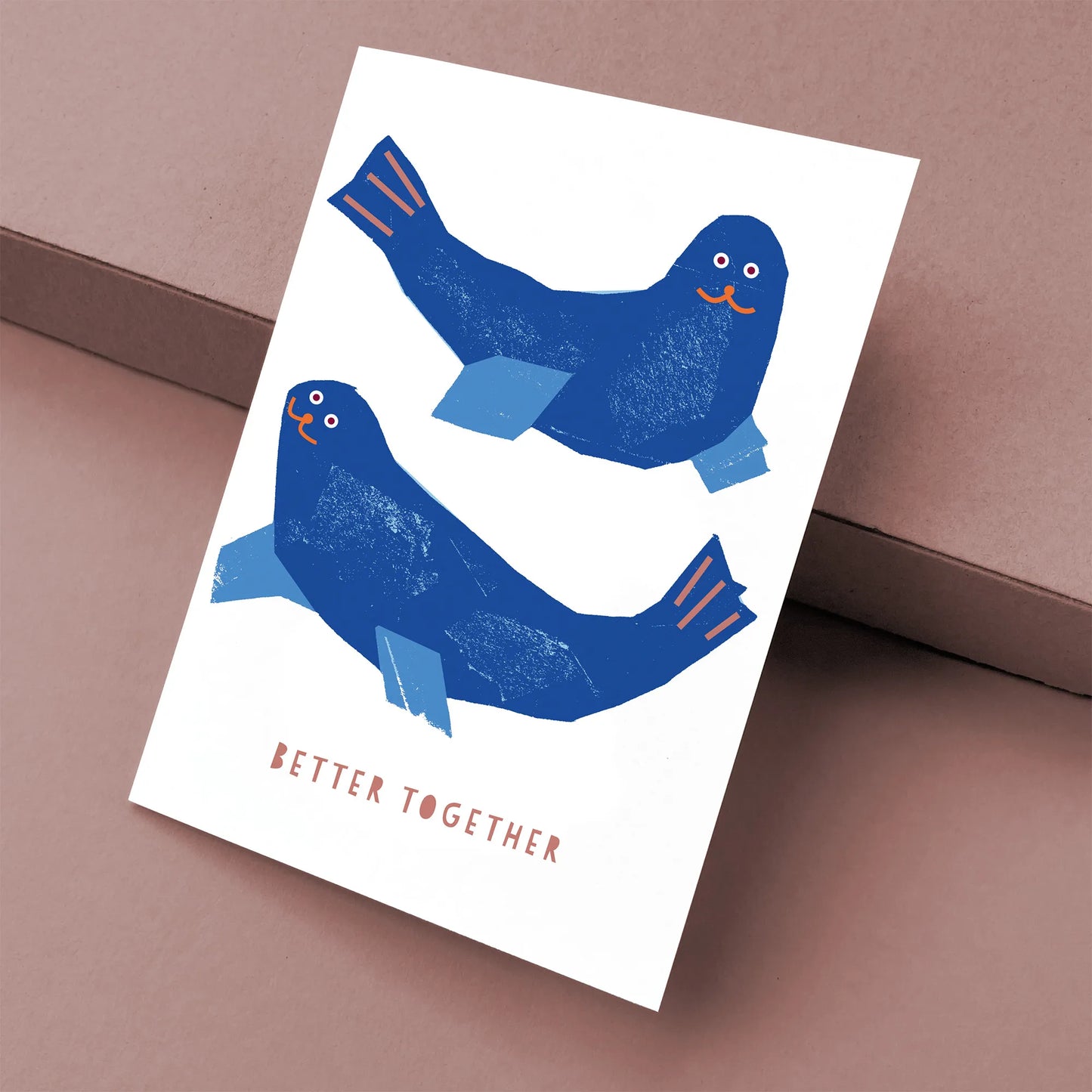 Kautzi greeting card featuring two playful blue seals with happy expressions and the text "Better Together," displayed at an angle with a brown kraft envelope.