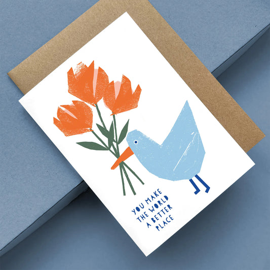 Kautzi greeting card with a light blue seagull holding bright orange flowers, with the text "You make the world a better place," displayed at an angle with a brown kraft envelope.