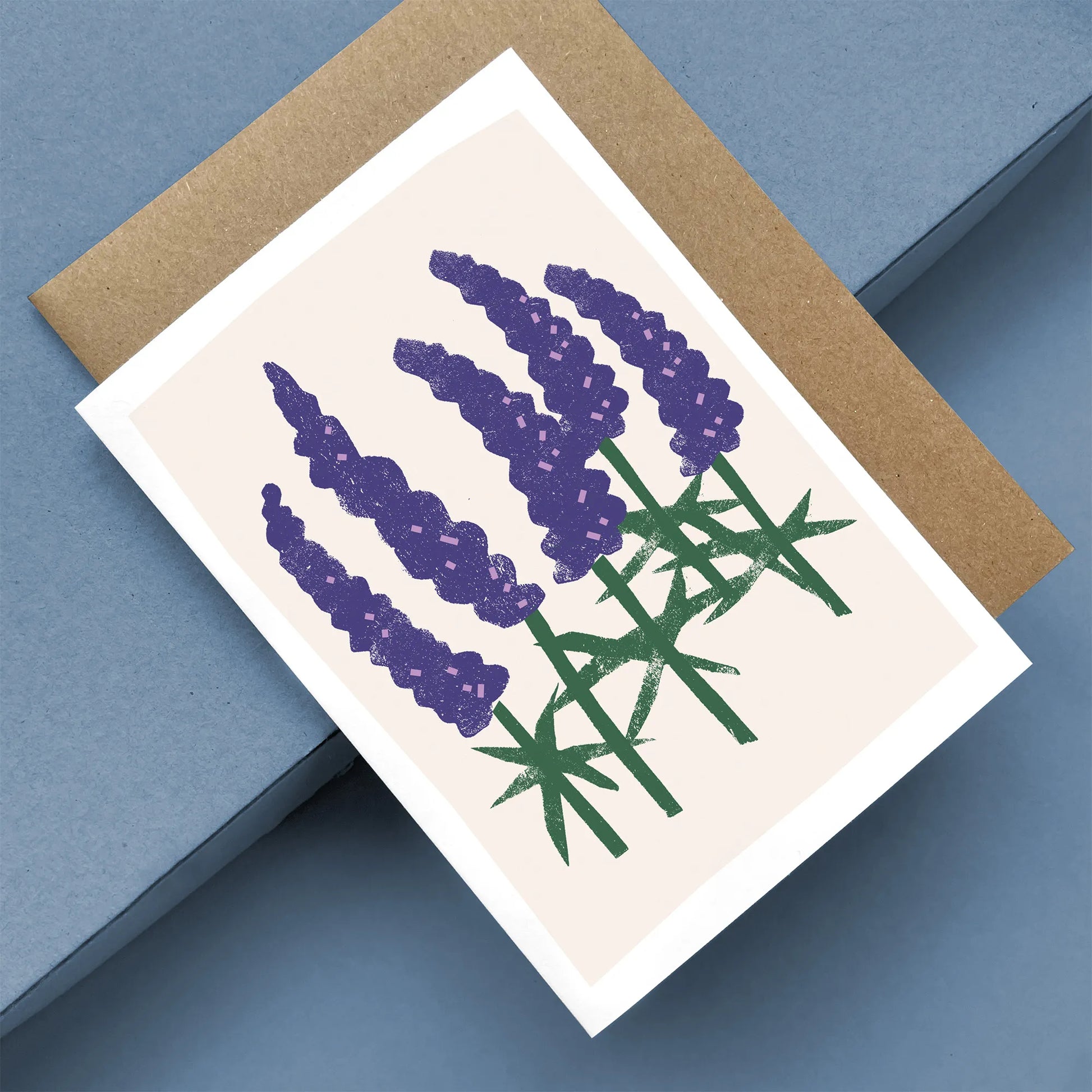 Kautzi Wild Lupines greeting card featuring bold purple lupine flowers with green stems on a soft pink background, displayed at an angle with a brown kraft envelope.