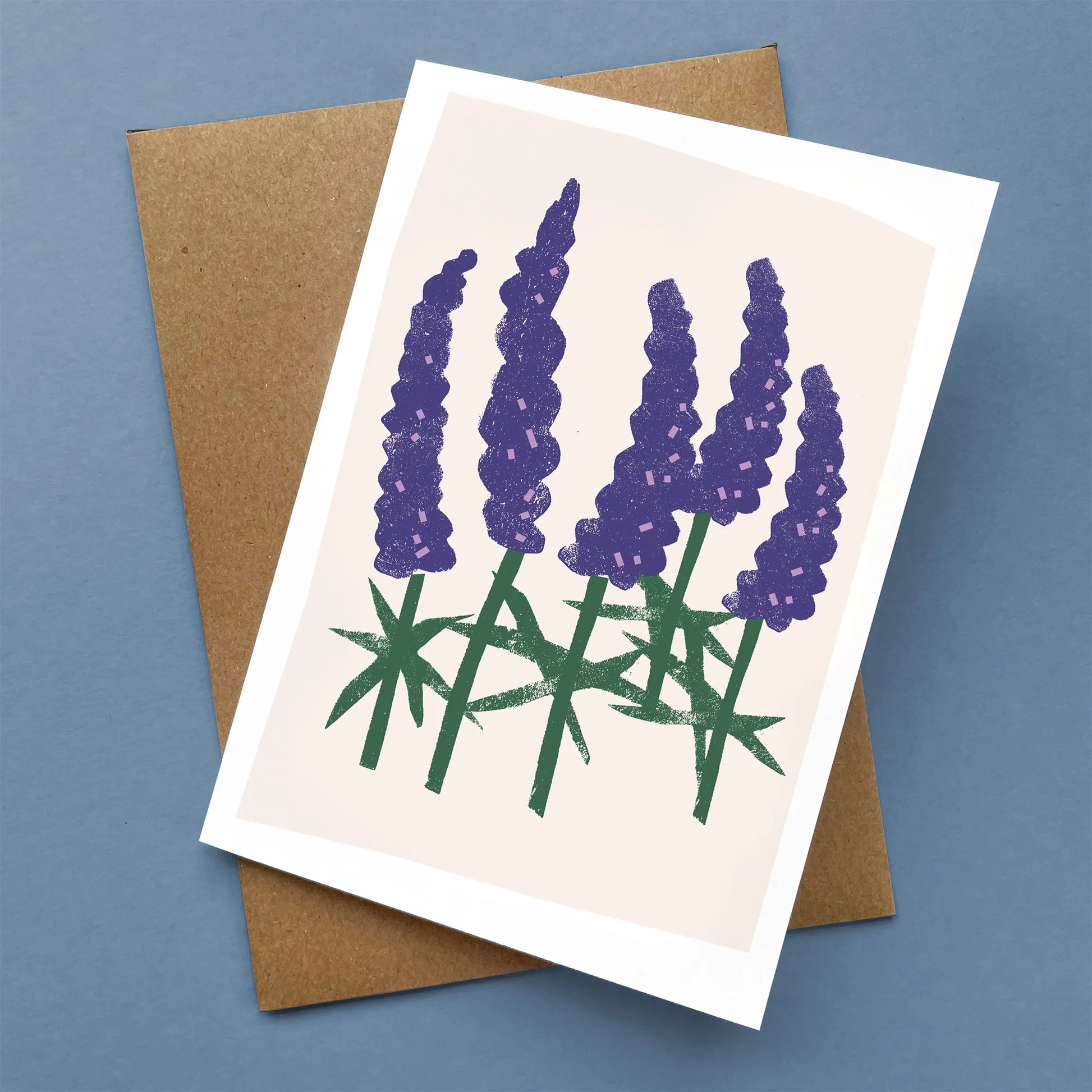 Kautzi Wild Lupines greeting card with hand-cut paper-style lupine flowers in purple and green on a soft pink background, shown flat with a brown kraft envelope.