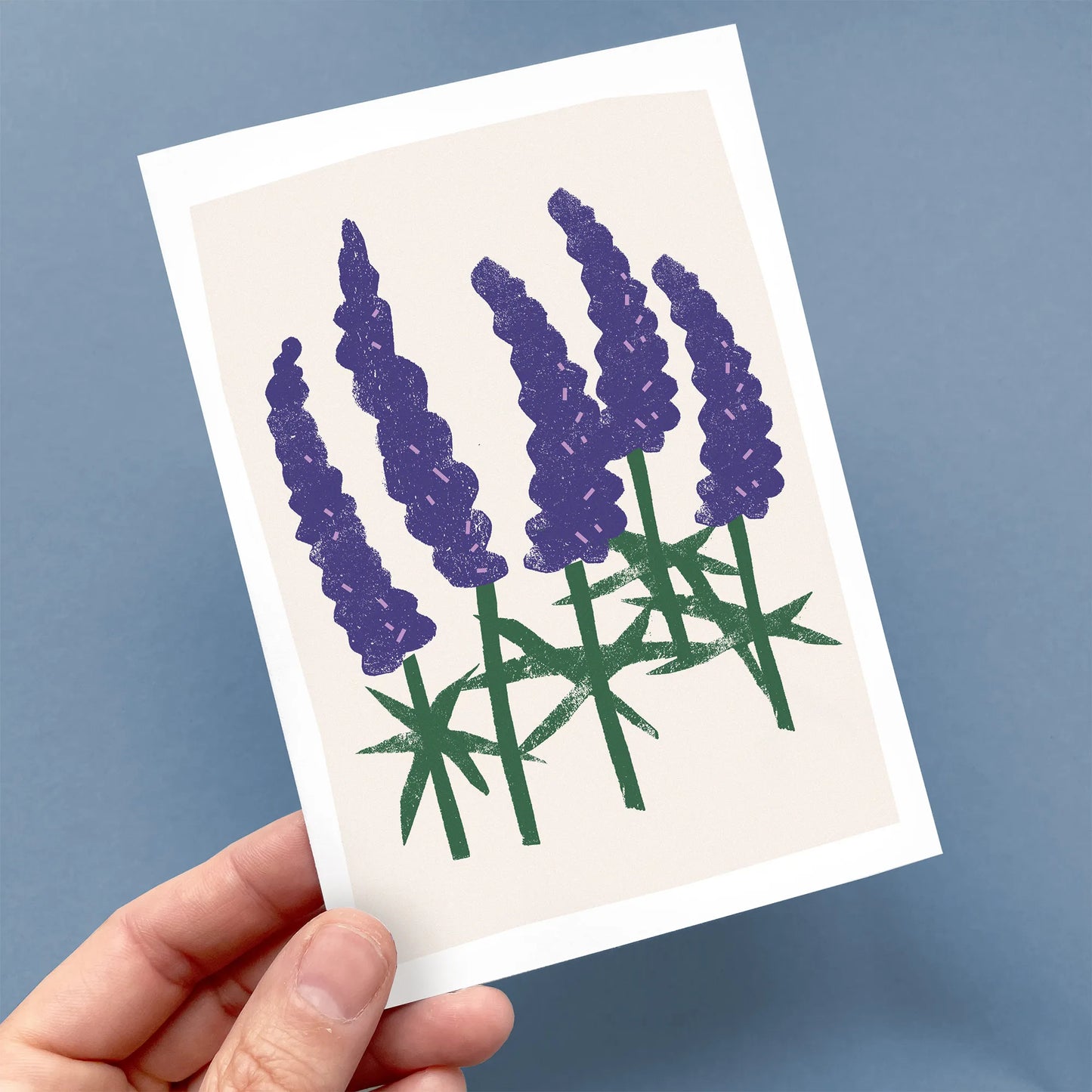 Kautzi Wild Lupines greeting card held in hand, featuring purple lupine flowers with green stems on a soft pink background.