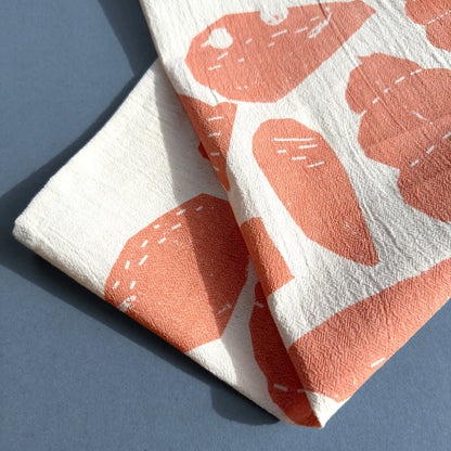 A close-up of the Beach Combings tea towel, highlighting the textured cotton fabric and the intricate details of the hand-cut shell illustrations in a warm coral shade. The screen-printing technique adds a slightly organic, imperfect touch.