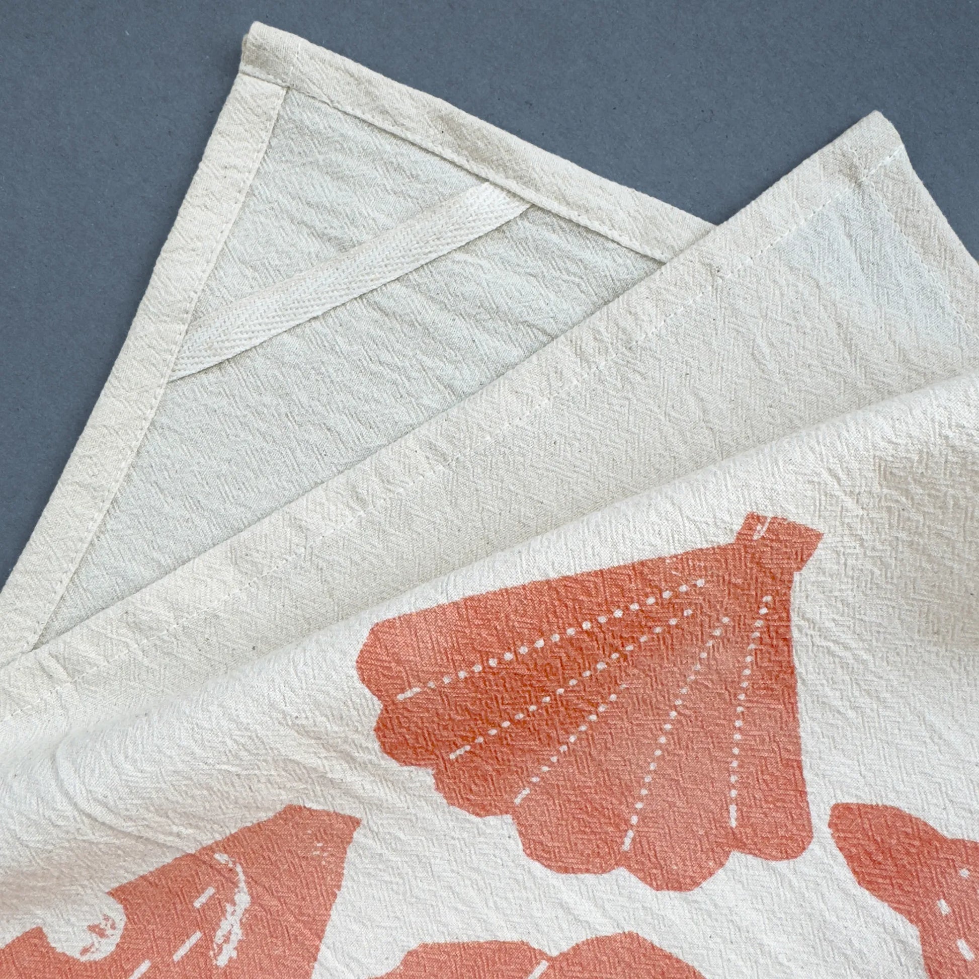 The Beach Combings tea towel, showing a sewn-in hanging loop on the back corner for easy storage. The fabric has a natural off-white base with a coral shell print and a subtle woven texture.