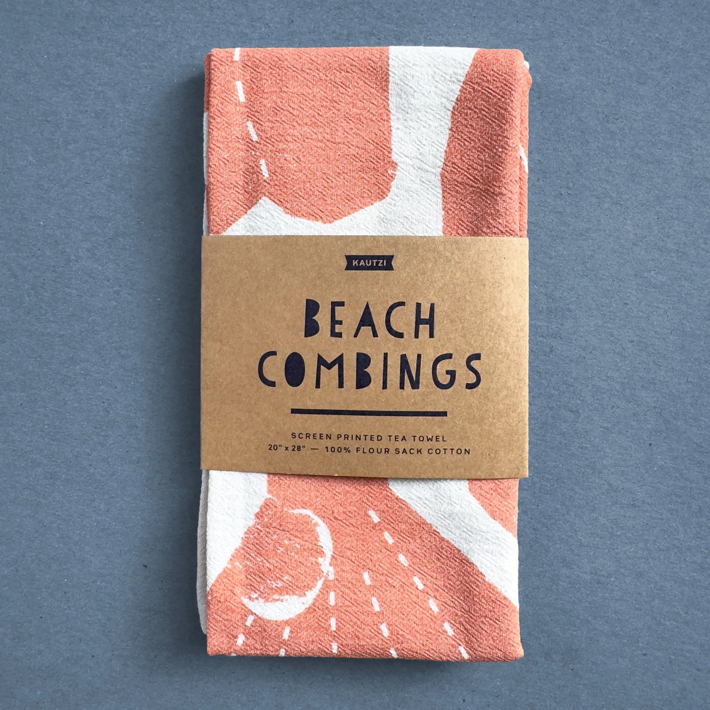 The Beach Combings tea towel by Kautzi, neatly folded and wrapped in a kraft paper label featuring the product name and details. The design showcases hand-cut seashell illustrations in a warm coral hue on natural white flour sack cotton.