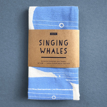 The Singing Whales tea towel by Kautzi, neatly folded and wrapped in kraft paper packaging with the product name and design details.