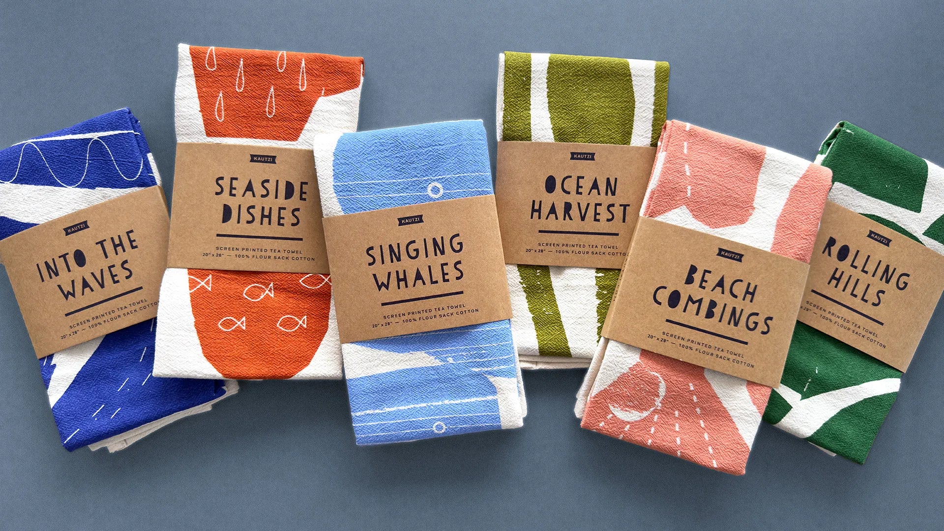 A collection of six screen-printed tea towels by Kautzi, neatly folded and wrapped in kraft paper labels. Each design showcases a unique coastal-inspired illustration in bold, nature-inspired colors: deep blue (Into the Waves), warm orange (Seaside Dishes), soft coral (Beach Combings), earthy green (Ocean Harvest), fresh green (Rolling Hills), and bright blue (Singing Whales). The towels are arranged on a muted blue background, highlighting their modern, minimal aesthetic and textured cotton fabric.