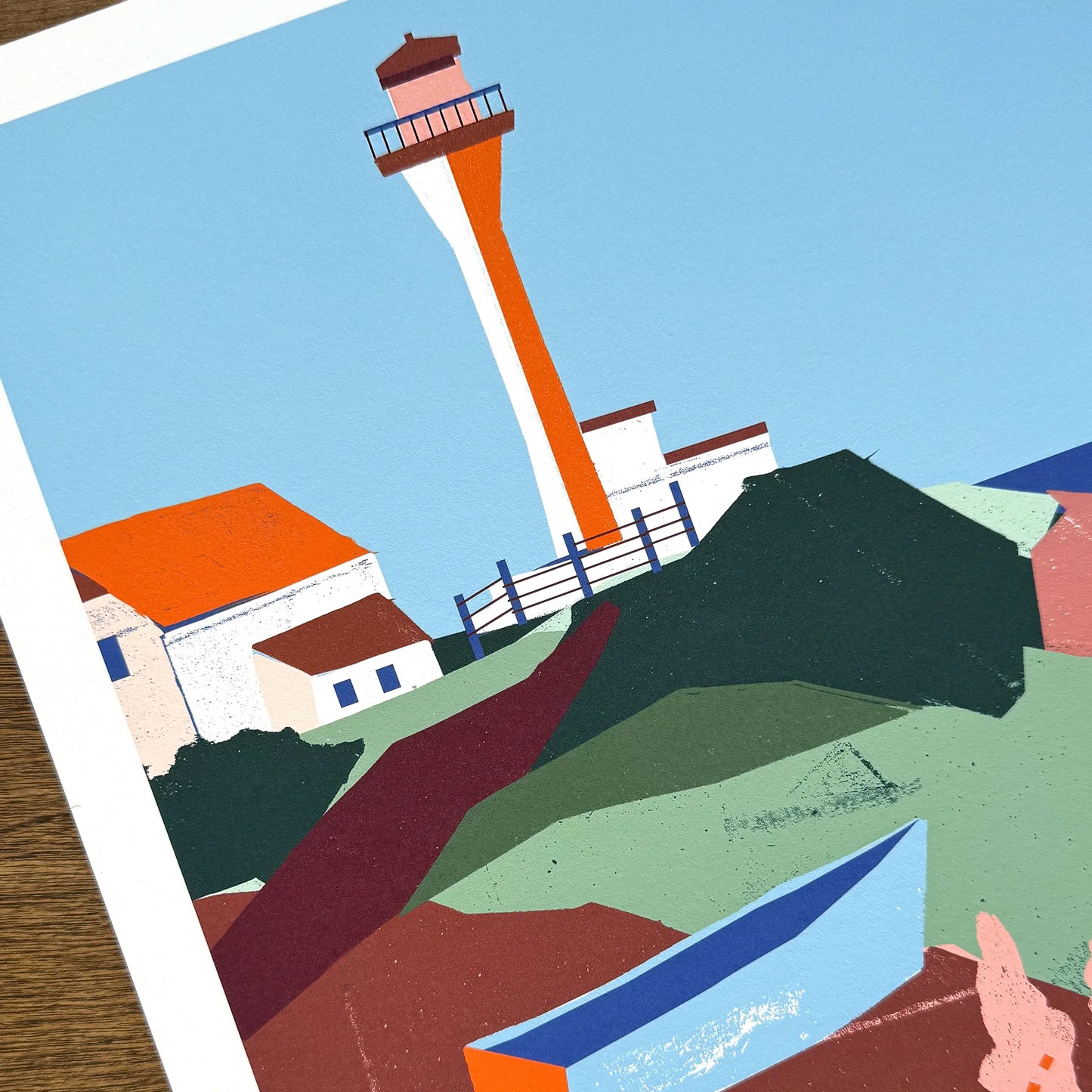 Close-up detail of Kautzi's Cape Forchu lighthouse art print, showing the bold collage-style illustration of the distinctive orange-and-white tower, keeper's buildings, and layered green and burgundy coastal rocks. Printed on archival cotton paper in Cape Breton, Nova Scotia, Canada.