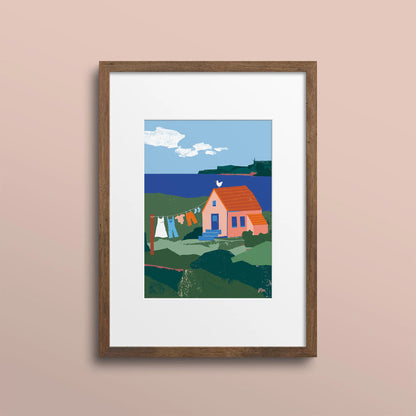 Kautzi Illustrations 5x7 Clothesline mini art print displayed in a wooden frame against a soft pink wall, showing a coastal house and clothesline with ocean and hills in the background, inspired by Cape Breton, Nova Scotia, Canada.