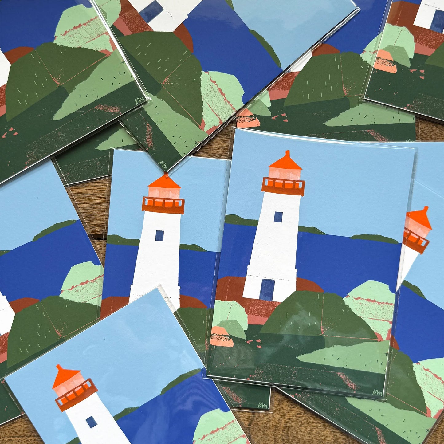 Multiple 5×7 art prints of the Louisbourg Lighthouse fanned out on a wooden surface, each sealed in a clear cello sleeve with a chipboard back.
