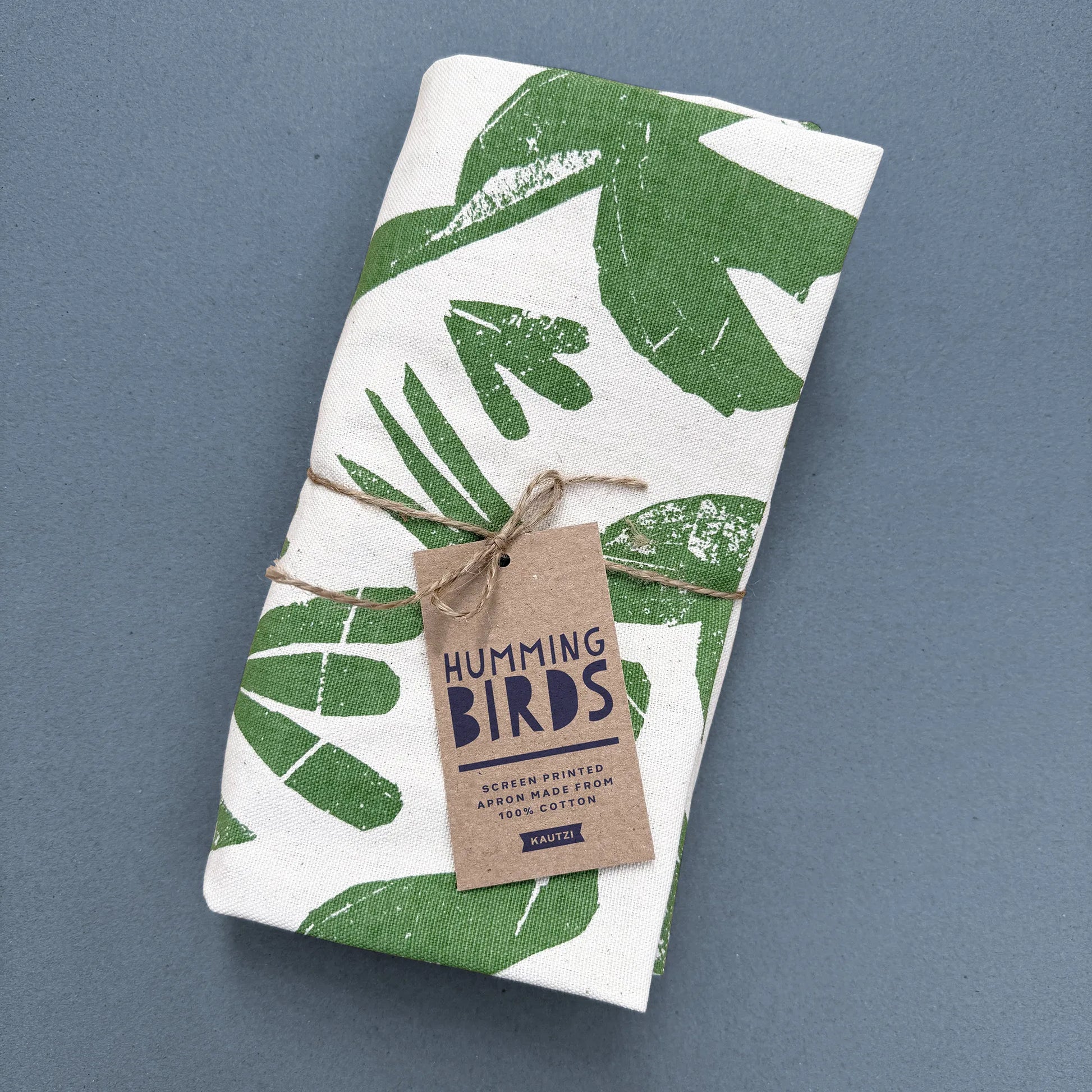Folded Kautzi Hummingbirds apron tied with twine and kraft hang tag, green screen-printed hummingbird design on natural cotton, photographed on a blue background, Cape Breton, Nova Scotia.