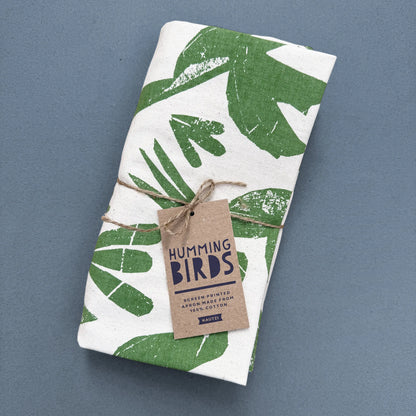 Folded Kautzi Hummingbirds apron tied with twine and kraft hang tag, green screen-printed hummingbird design on natural cotton, photographed on a blue background, Cape Breton, Nova Scotia.