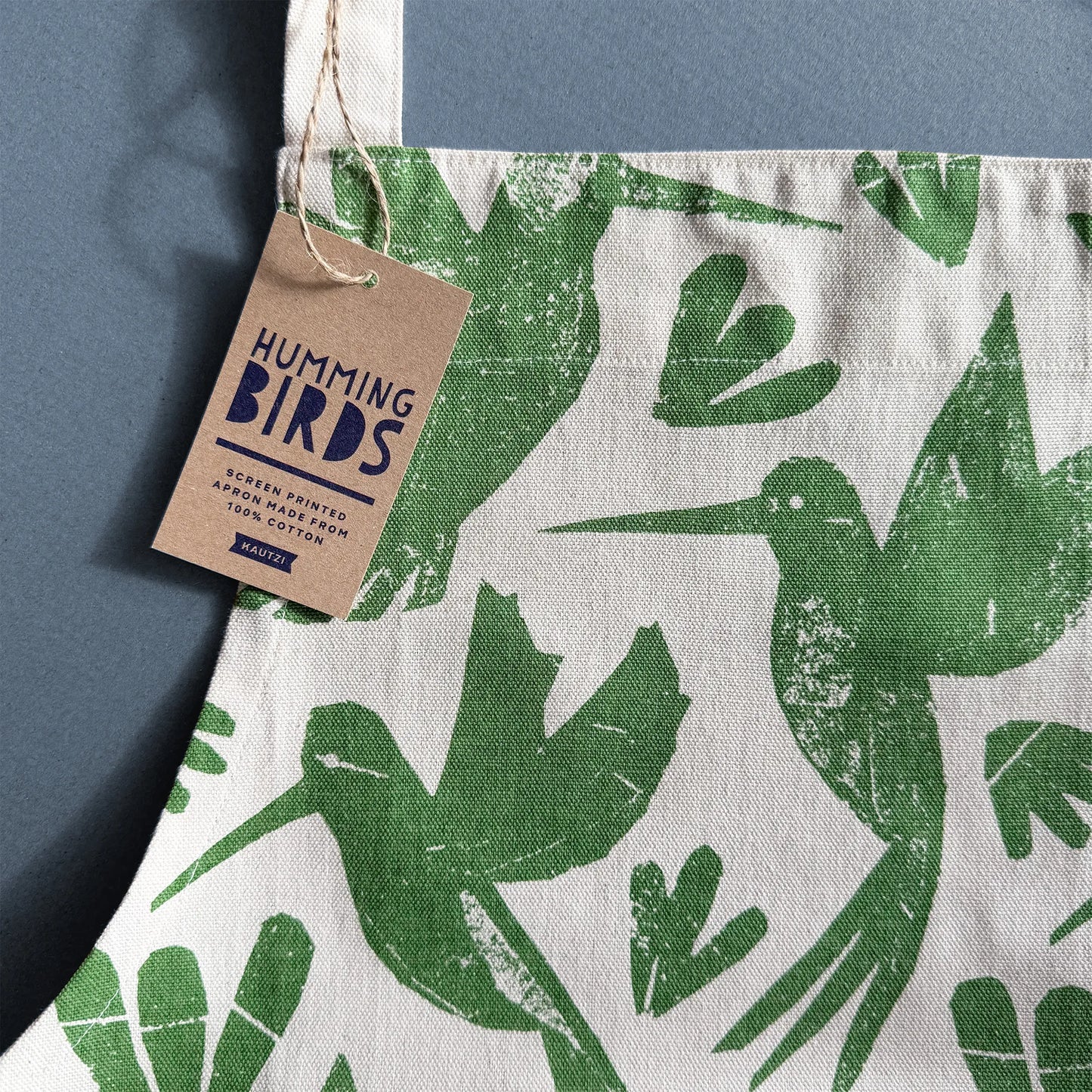 Close-up of Kautzi Hummingbirds apron showing green hummingbird print on cotton fabric with kraft hang tag on a blue background, illustrated textile from Nova Scotia, Canada.