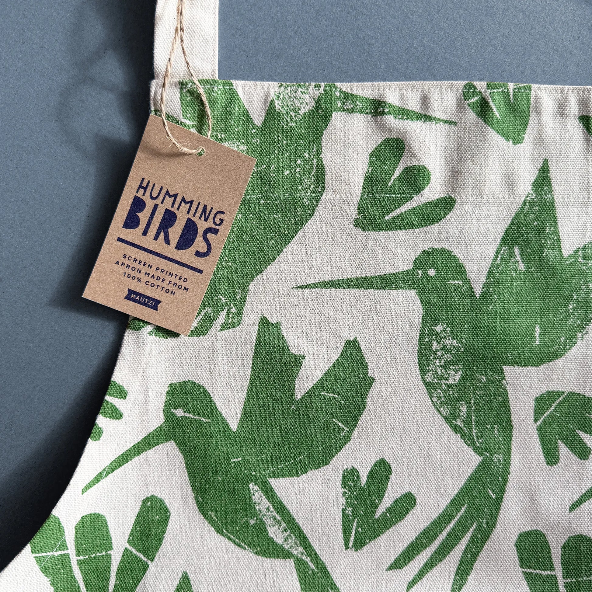 Close-up of Kautzi Hummingbirds apron showing green hummingbird print on cotton fabric with kraft hang tag on a blue background, illustrated textile from Nova Scotia, Canada.