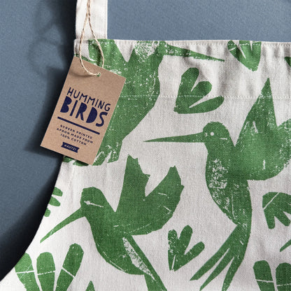Close-up of Kautzi Hummingbirds apron showing green hummingbird print on cotton fabric with kraft hang tag on a blue background, illustrated textile from Nova Scotia, Canada.