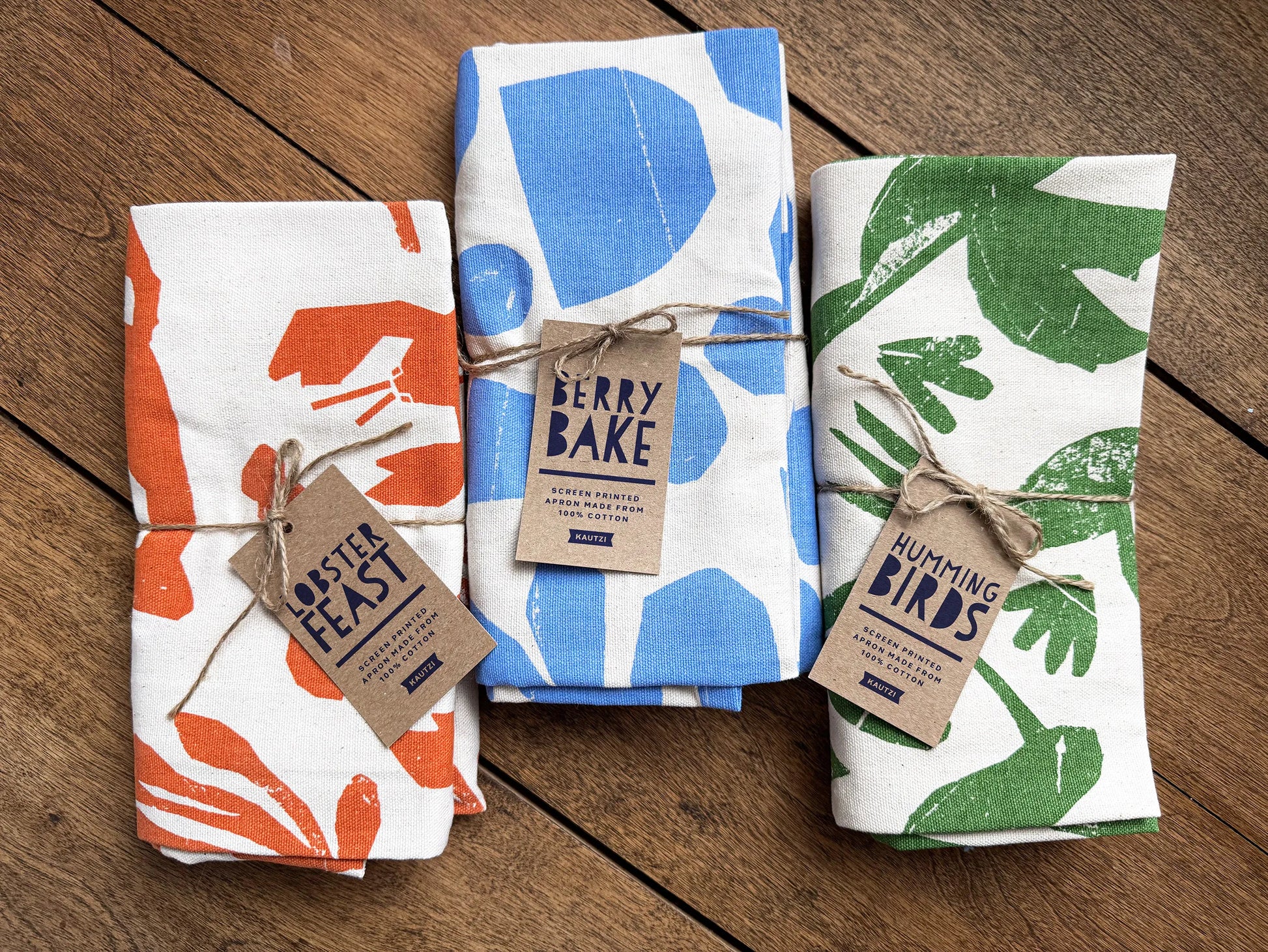 Three folded Kautzi aprons side by side on a wooden surface, including the green Hummingbirds design alongside Berry Bake and Lobster Feast, screen printed cotton aprons from Cape Breton, Nova Scotia, Canada.