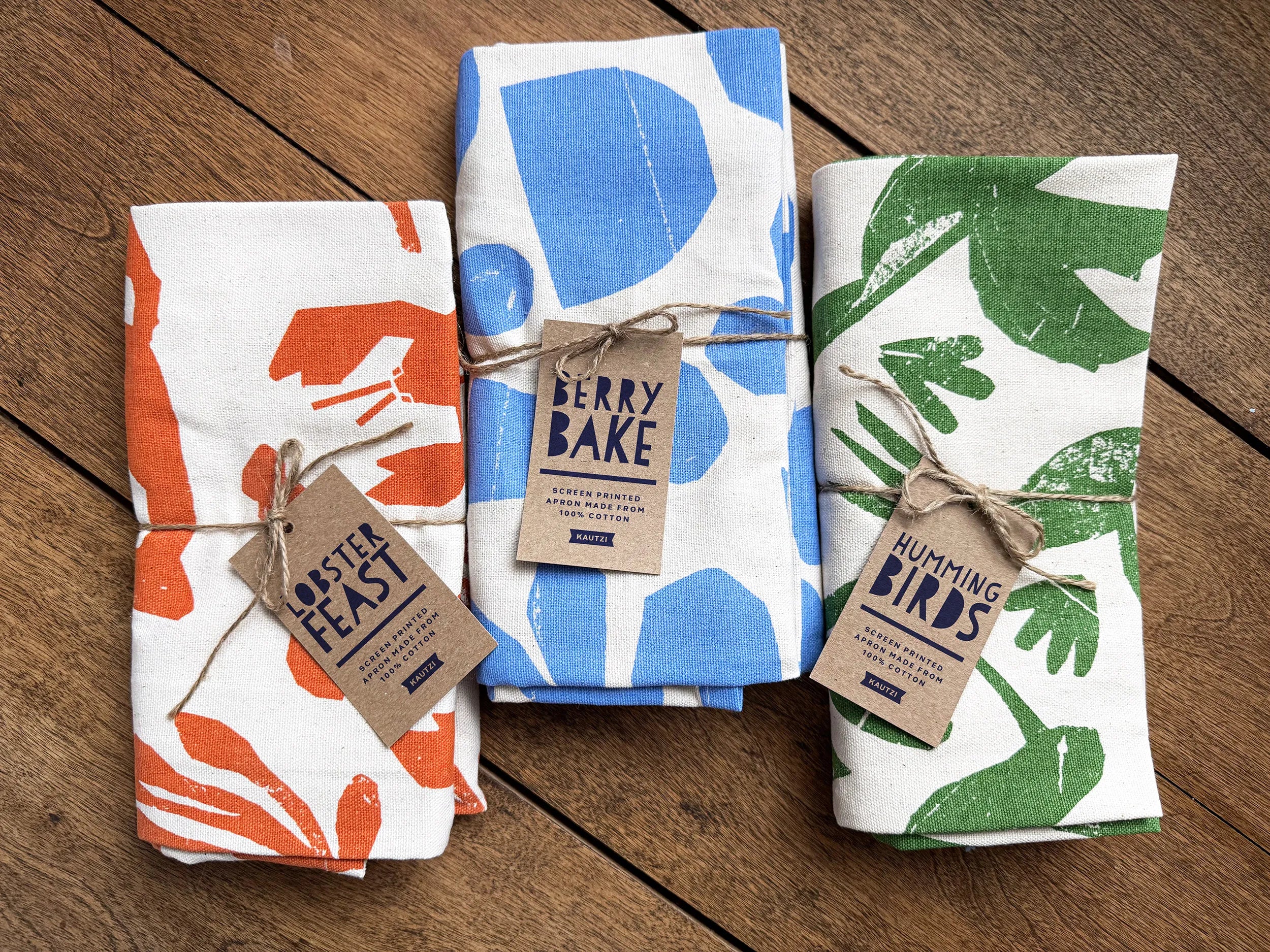 Three folded Kautzi aprons side by side on a wooden surface, including the green Hummingbirds design alongside Berry Bake and Lobster Feast, screen printed cotton aprons from Cape Breton, Nova Scotia, Canada.
