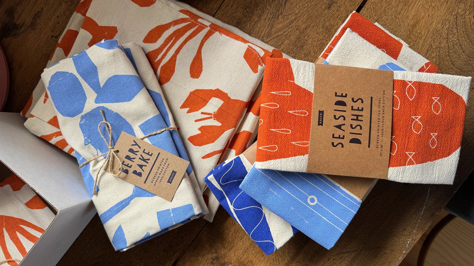 Kautzi screen-printed tea towels and aprons in coastal colours, folded and packaged with kraft labels, designed in Cape Breton, Nova Scotia