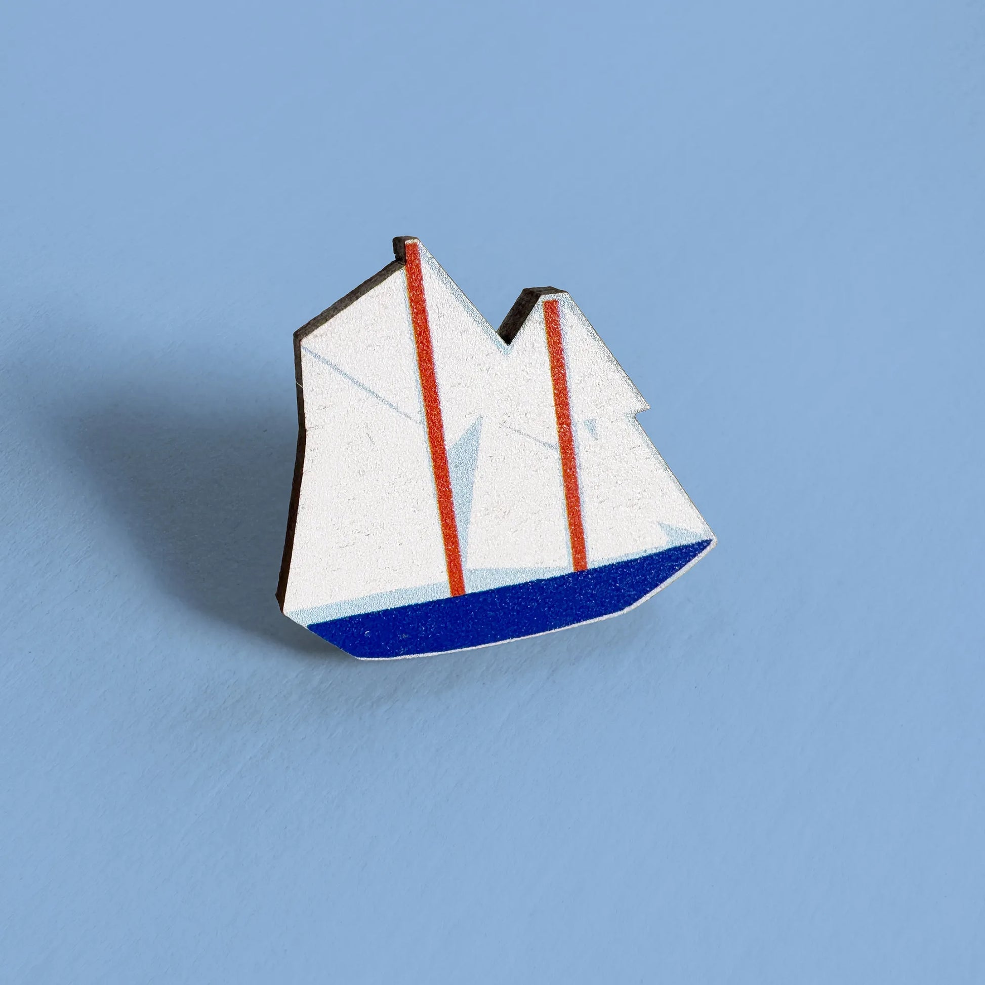 Close-up of Kautzi maple wood veneer boat pin badge with white sails, red masts, and blue hull, inspired by Nova Scotia’s Bluenose