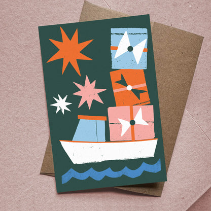 Front of Christmas card titled "Christmas Cargo" showing a lobster boat stacked with gifts and stars, with kraft envelope beneath.