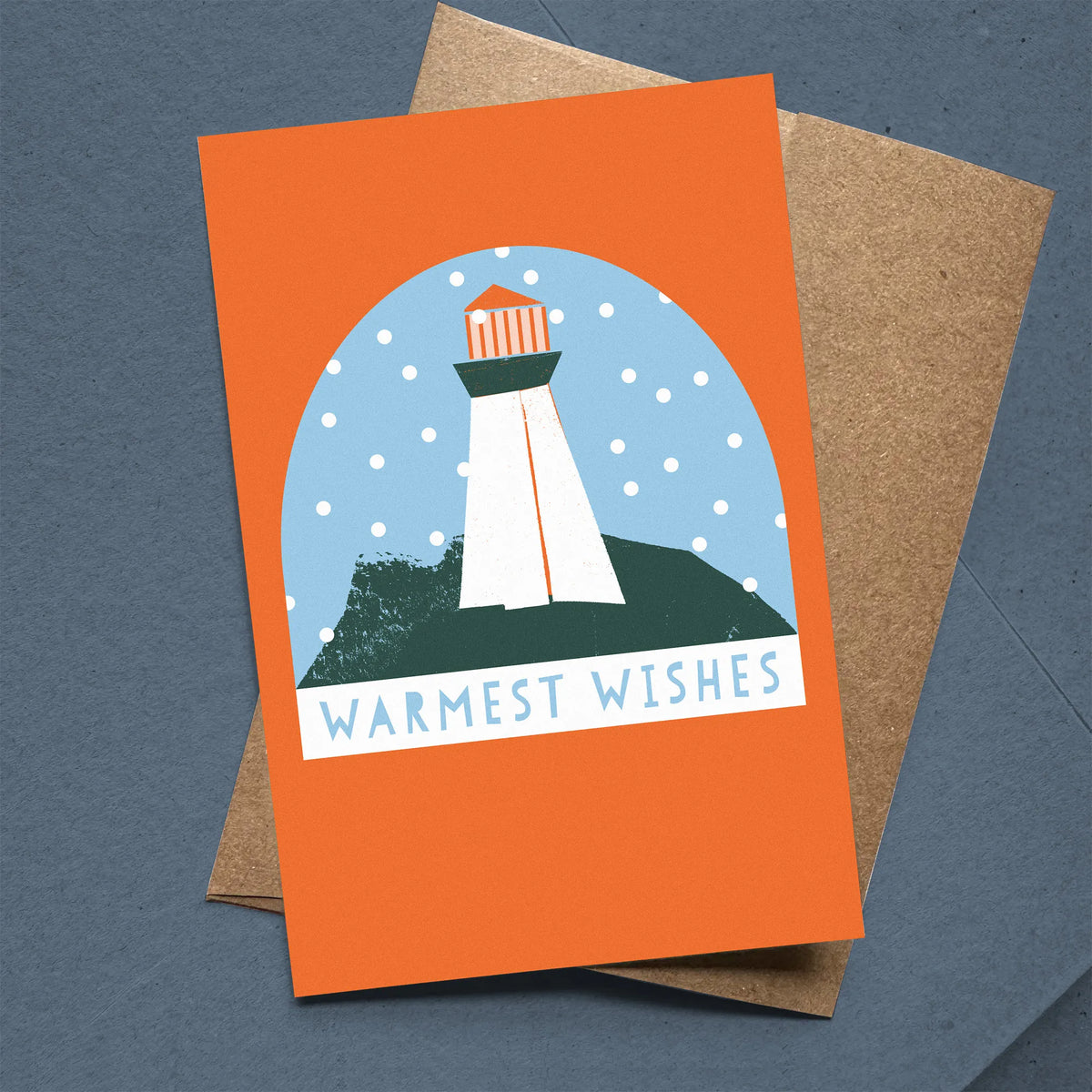 Kautzi "Warmest Wishes" Christmas card with a Maritime lighthouse snow globe illustration in orange, teal, and blue, leaning against a kraft envelope on a dark slate background. Nova Scotia coastal holiday greeting card