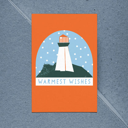 Kautzi Coastal Christmas holiday card featuring an illustrated lighthouse in a snow globe scene on a bold orange background, reading "Warmest Wishes," shown with a kraft paper envelope on a blue background. 