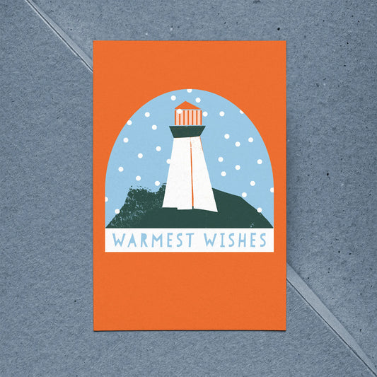 Kautzi Coastal Christmas holiday card featuring an illustrated lighthouse in a snow globe scene on a bold orange background, reading "Warmest Wishes," shown with a kraft paper envelope on a blue background. 