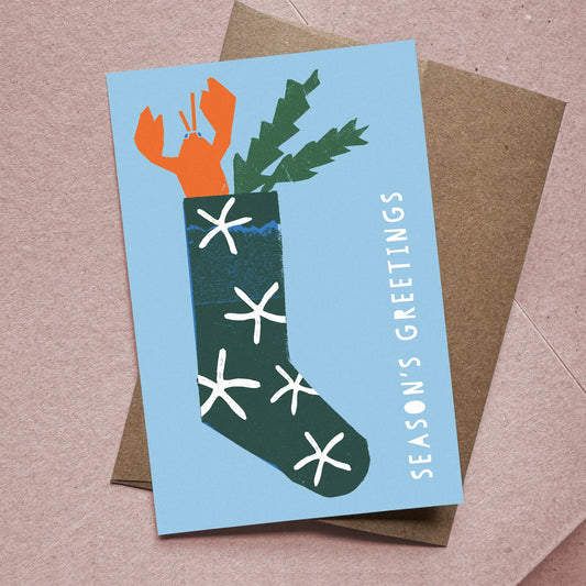 Flat Christmas card with lobster-in-stocking illustration and kraft envelope on soft pink background.
