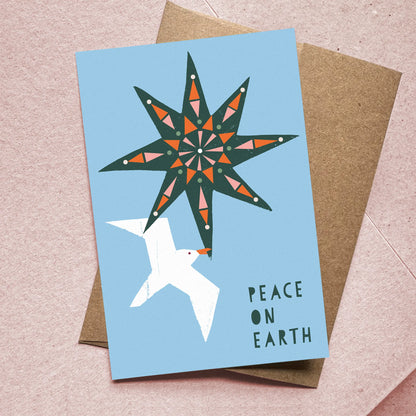 Kautzi Peace on Earth Christmas card featuring a bold illustrated eight-pointed star and white seagull on a sky blue background, shown overlapping a kraft paper envelope on a pink textured surface. Canadian holiday greeting card designed in Nova Scotia