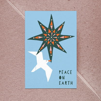 Kautzi Peace on Earth greeting card with a modern illustrated geometric star in teal, orange, and pink above a white seagull in flight, reading "Peace on Earth," shown with a kraft envelope on a light pink background. Atlantic Canada Christmas card