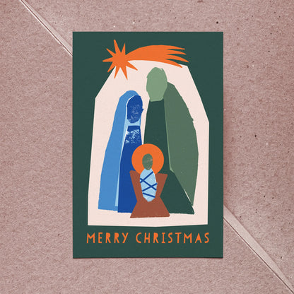 Kautzi Silent Night Christmas card featuring a modern illustrated nativity scene with bold teal, blue, and orange tones on a deep green background, reading "Merry Christmas," shown with a kraft paper envelope on a light pink background — Canadian holiday greeting card