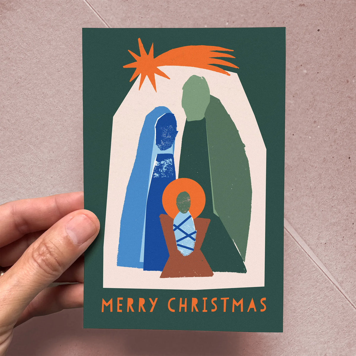 Hand holding the Kautzi Silent Night "Merry Christmas" greeting card, showing the full front design of a modern illustrated nativity scene in teal, blue, and orange on a deep green background. Atlantic Canada Christmas card, designed in Nova Scotia
