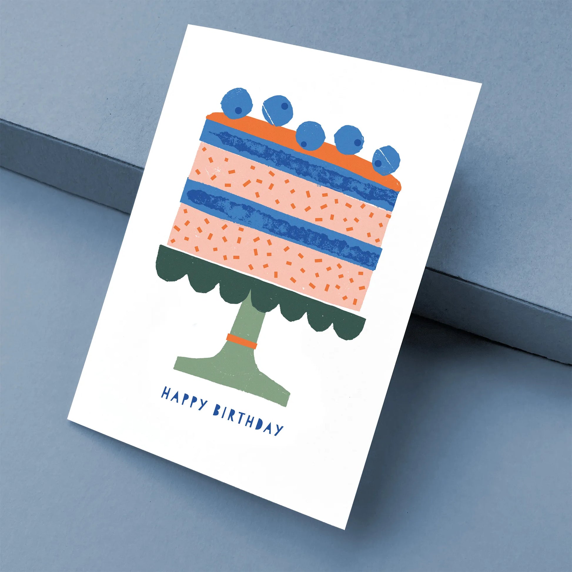 Kautzi greeting card featuring a colourful birthday cake illustration with blueberries on top, shown at an angle on a blue background – designed in Cape Breton, Nova Scotia.