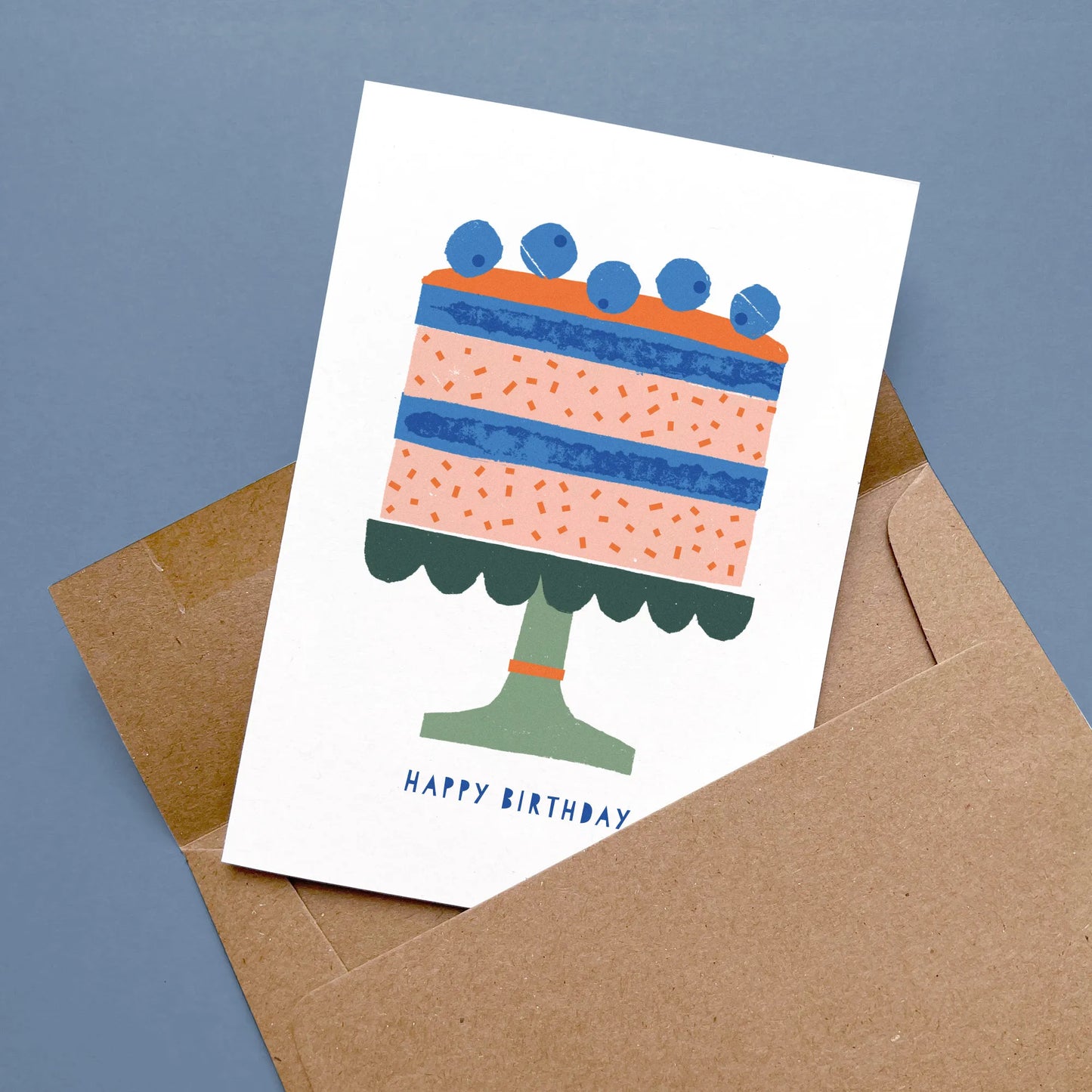 Kautzi birthday card with a modern birthday cake illustration in shades of pink, blue, and green, displayed flat with a kraft envelope on a blue background – designed in Cape Breton, Nova Scotia.