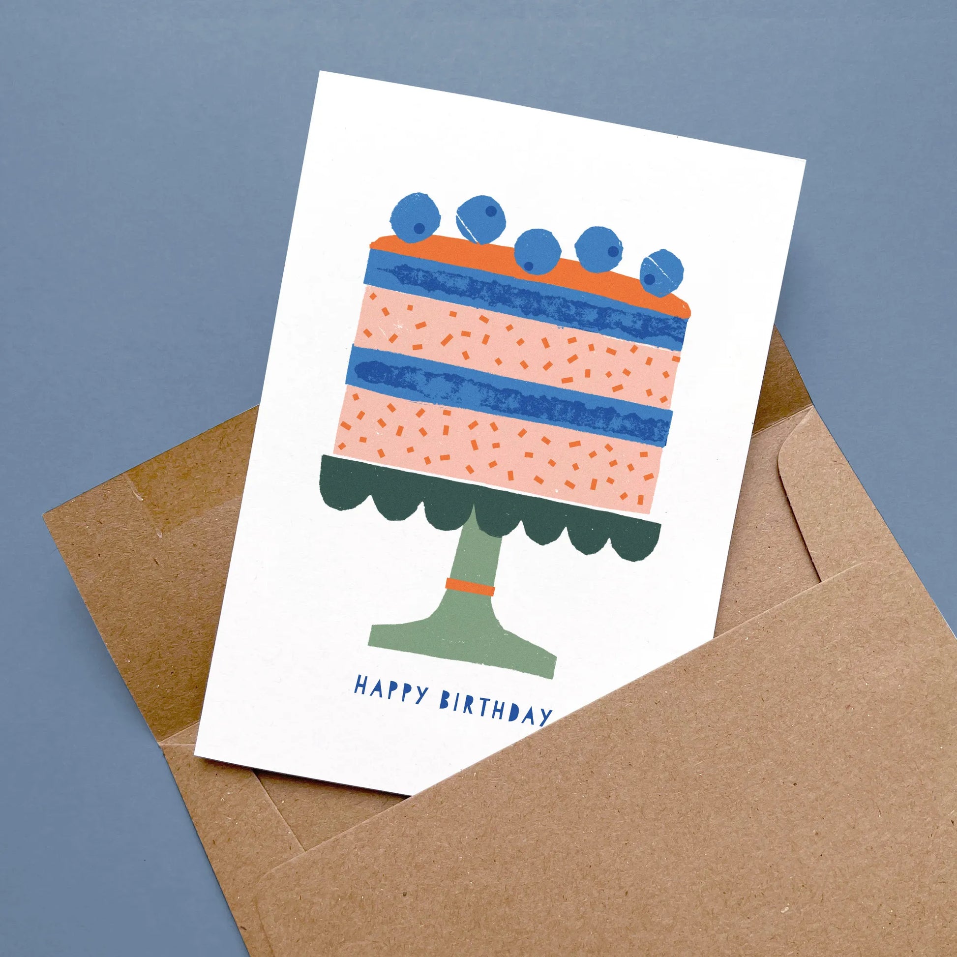 Kautzi birthday card with a modern birthday cake illustration in shades of pink, blue, and green, displayed flat with a kraft envelope on a blue background – designed in Cape Breton, Nova Scotia.